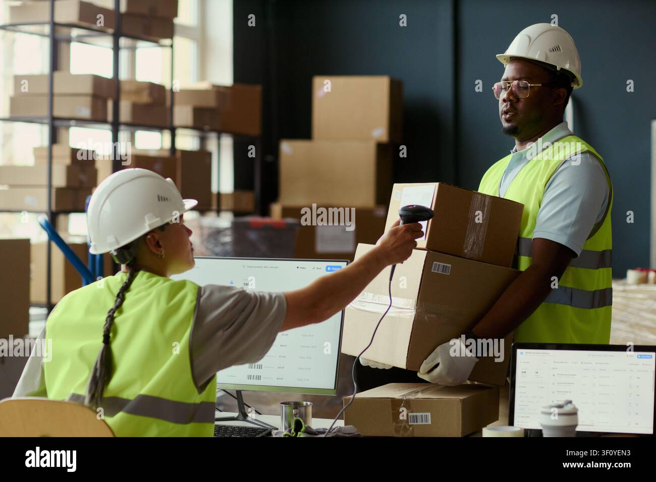 Lavoratore di magazzino afroamericano che trasporta scatole mentre una collega scansiona il codice a barre alla stazione di imballaggio, mostrando il controllo dell'inventario e il flusso logistico. Per il marketing della catena di fornitura Foto Stock