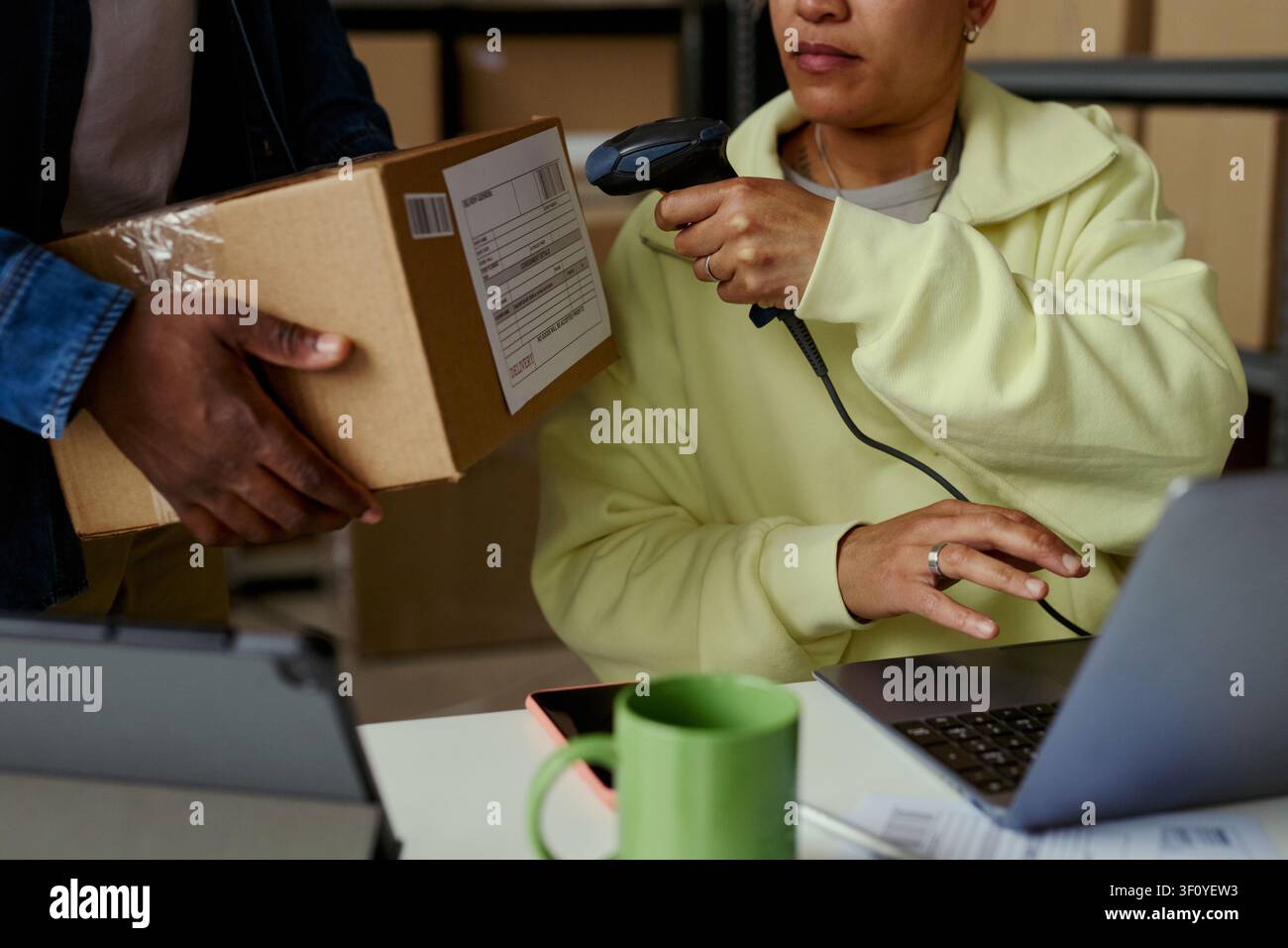Donna biracial di mezza età che scansiona l'etichetta di spedizione su un pacco mentre un collega tiene una scatola vicino al laptop, elaborando l'adempimento dell'ordine. Ideale per e-commerce, flusso di magazzino, servizio di consegna Foto Stock