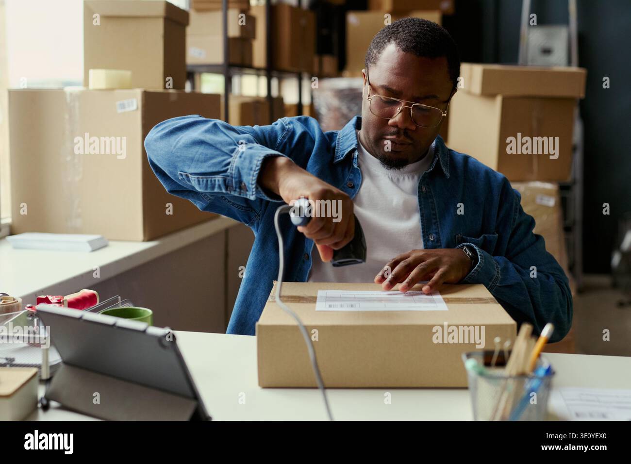 Scatto medio di un giovane uomo afroamericano che scansiona l'etichetta di spedizione su un pacco, elaborando l'adempimento degli ordini in un ambiente di lavoro del magazzino. Ideale per e-commerce, logistica, marketing per piccole imprese Foto Stock