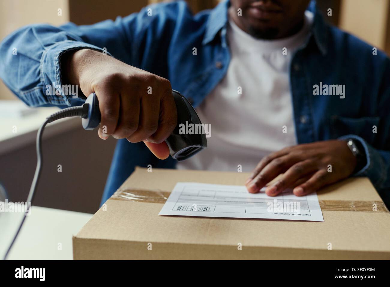 Primo piano di un giovane uomo nero che scansiona un codice a barre sull'etichetta di spedizione sopra un pacco, elaborando l'ordine per la consegna. Ideale per e-commerce, logistica di magazzino, marketing di adempimento Foto Stock