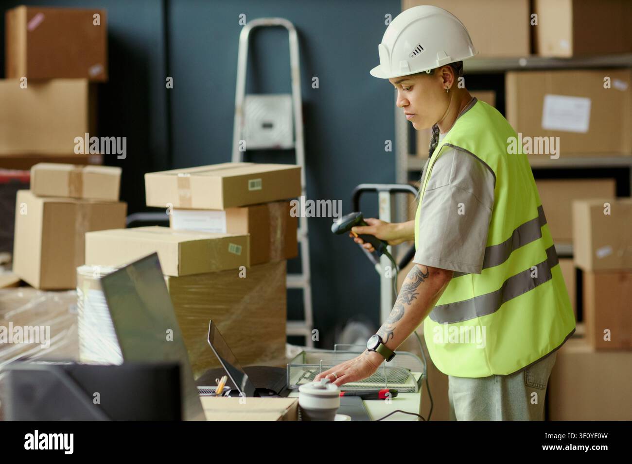 Giovane donna adulta biracial impiegata in magazzino che scansiona il codice a barre del pacco e controlla il laptop durante l'elaborazione dell'inventario nel centro di distribuzione. Ideale per logistica, spedizione, marketing della catena di fornitura Foto Stock