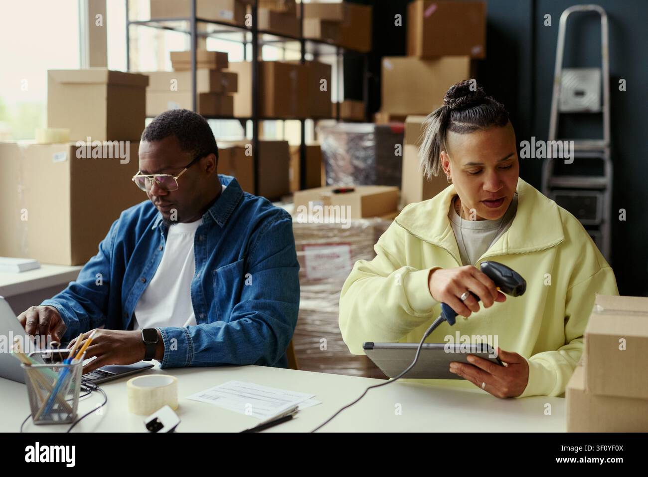 Uomo afroamericano che digita su un laptop mentre una giovane donna scansiona i dati dei pacchi su un tablet nell'ufficio del magazzino. Ideale per la gestione degli ordini e-commerce, la gestione delle scorte, le piccole imprese Foto Stock