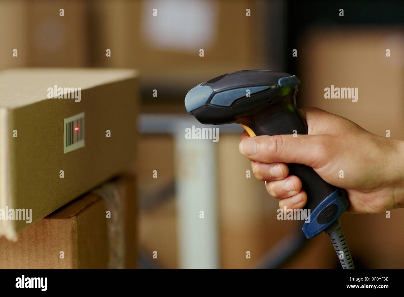 Primo piano di una mano adulta che scansiona un codice a barre su una scatola di cartone con un lettore portatile, gestendo l'inventario e il tracciamento degli ordini in magazzino. Ideale per la logistica, la vendita al dettaglio, il contenuto di spedizione Foto Stock