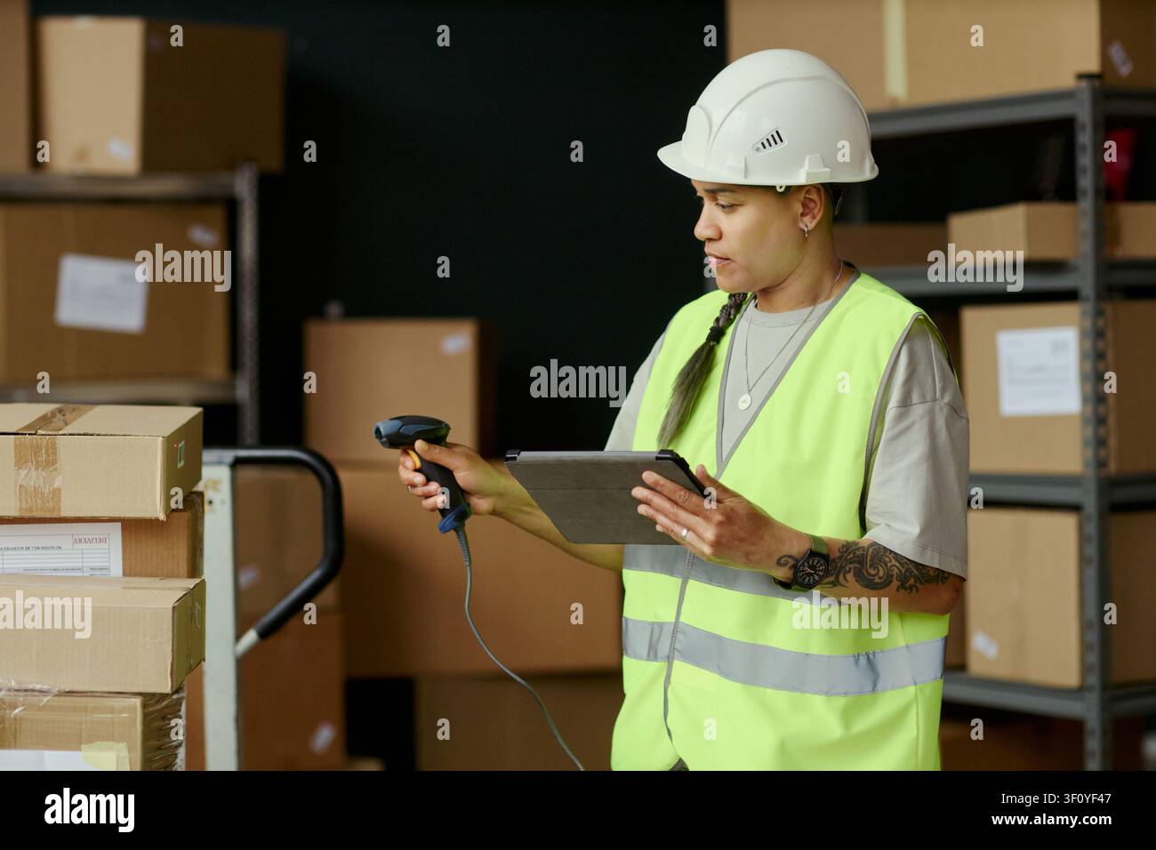 Giovane donna adulta birazziale addetta al magazzino che scansiona pacchi e controlla un tablet durante il controllo delle scorte, supportando le operazioni logistiche. Utile per la catena di fornitura, l'adempimento, la vendita al dettaglio Foto Stock