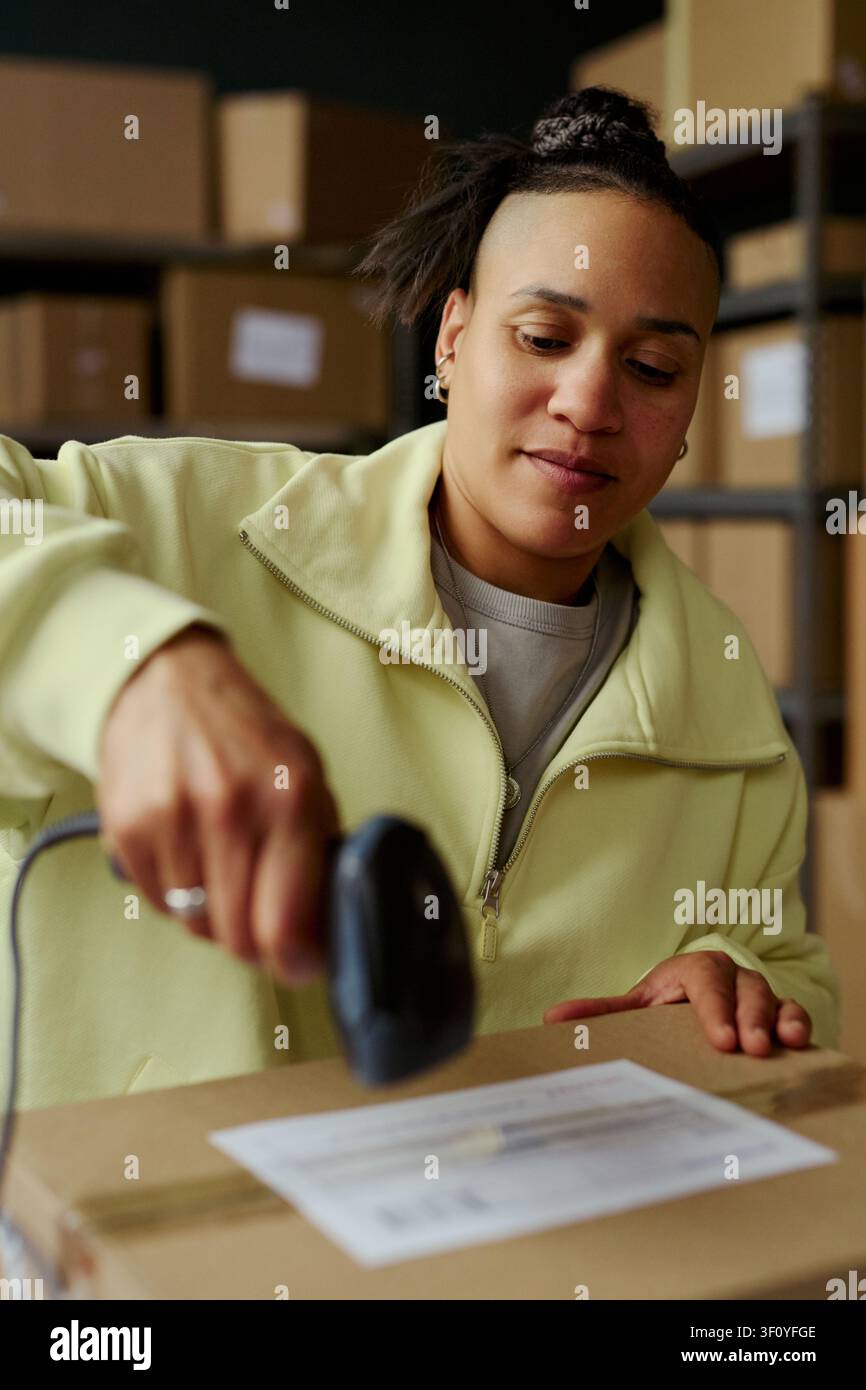 Scatto verticale di una giovane donna biracial che scansiona l'etichetta di spedizione su un pacco in un magazzino, elaborando l'adempimento di un ordine di vendita al dettaglio online. Ideale per e-commerce, logistica, marketing per piccole imprese Foto Stock