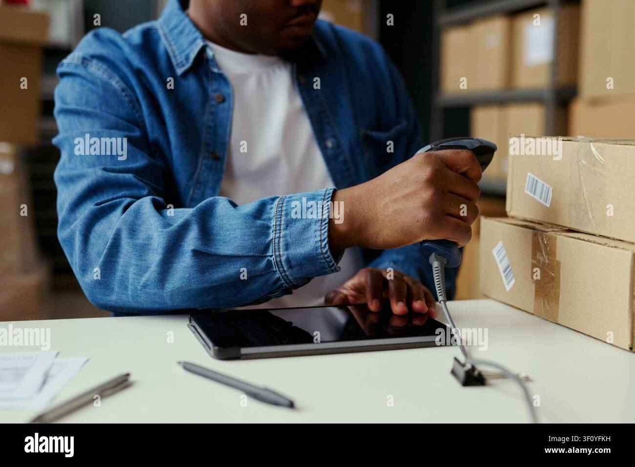 Primo piano di un giovane uomo nero che scansiona i codici a barre dei pacchi accanto a un tablet, elaborando l'inventario in un magazzino. Ideale per la gestione dell'e-commerce, la logistica, il tracciamento degli ordini Foto Stock