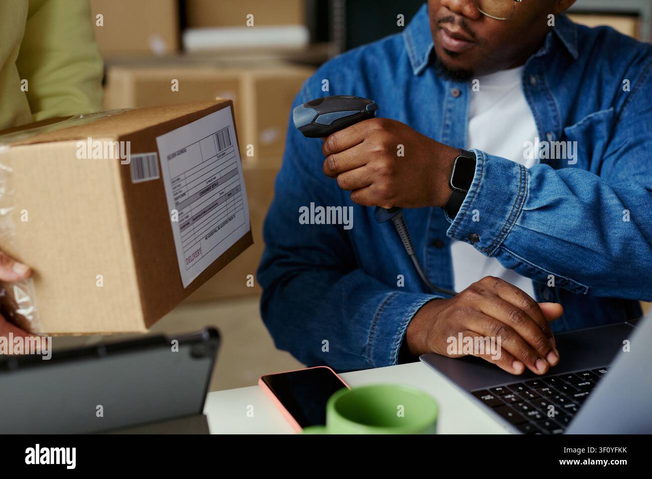 Primo piano di un giovane uomo nero che scansiona l'etichetta di spedizione su un pacco mentre inserisce i dati dell'ordine su un laptop, gestendo il flusso di lavoro di adempimento. Ideale per il marketing logistico dell'e-commerce Foto Stock