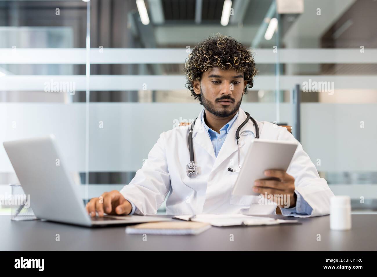Giovane medico maschio con capelli ricci che indossa un camice bianco e uno stetoscopio, lavora su un laptop e controlla i dati dei pazienti su un tablet digitale in un Foto Stock
