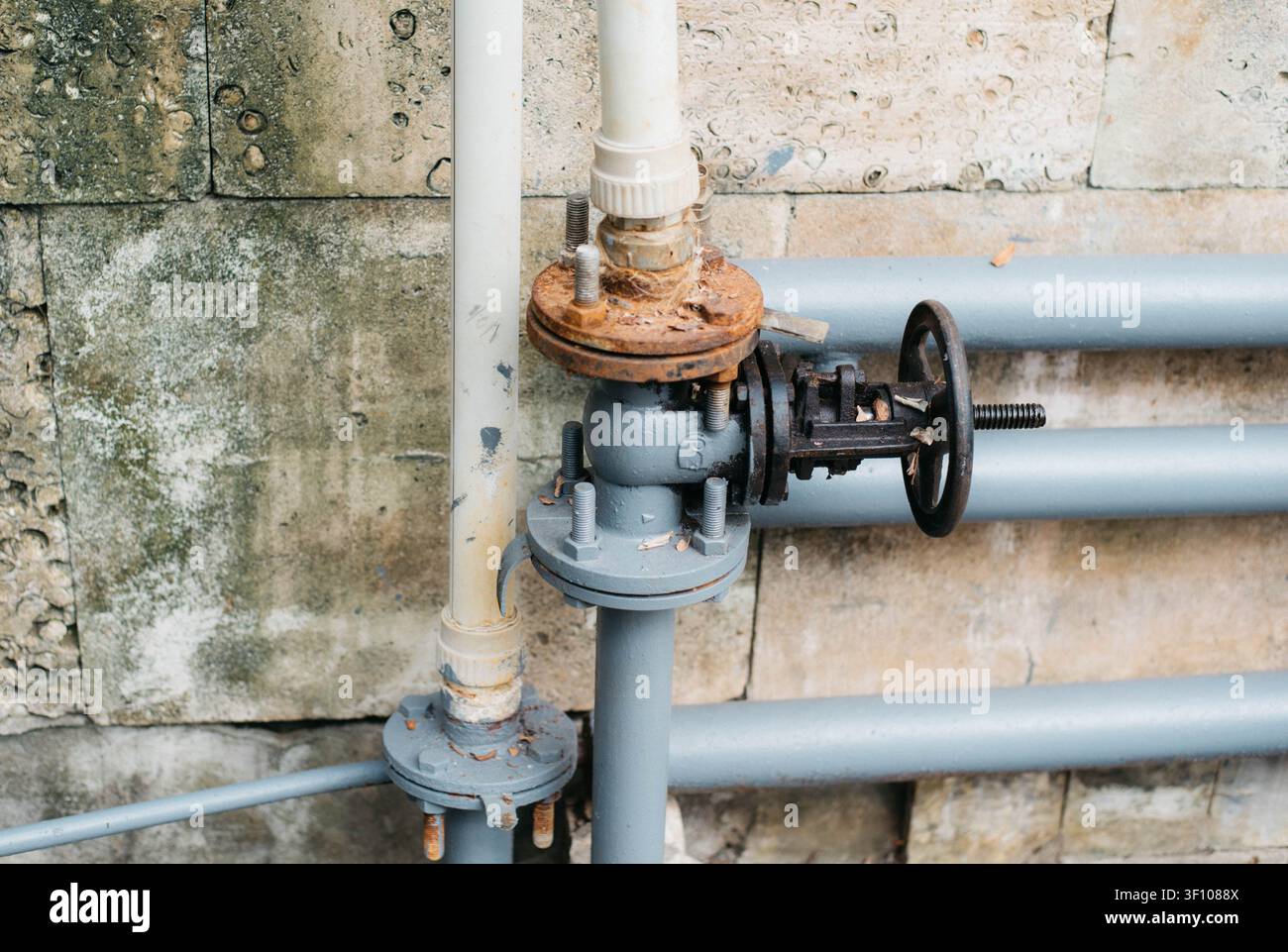 Primo piano di una pipeline industriale con una valvola metallica, flangia arrugginita e tubi in PVC montati su un muro di cemento consumato. Concetto di approvvigionamento idrico, idraulica, infrastrutture di servizi pubblici, manutenzione industriale, ingegneria e sistemi di pipeline. Foto Stock