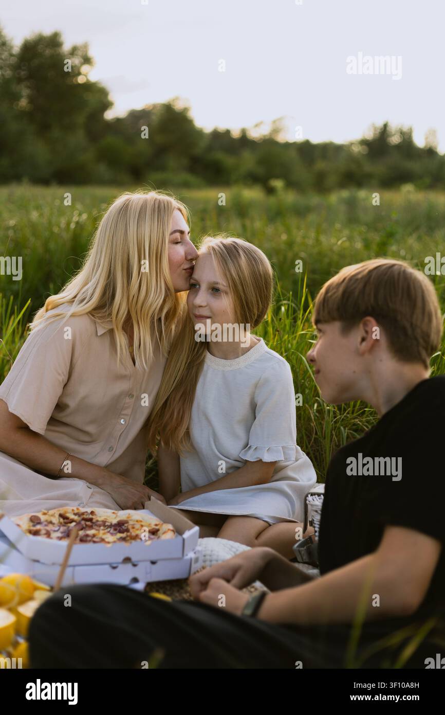 Una madre amorevole condivide un momento tenero con sua figlia mentre gode di un picnic con suo figlio in un prato illuminato dal sole Foto Stock