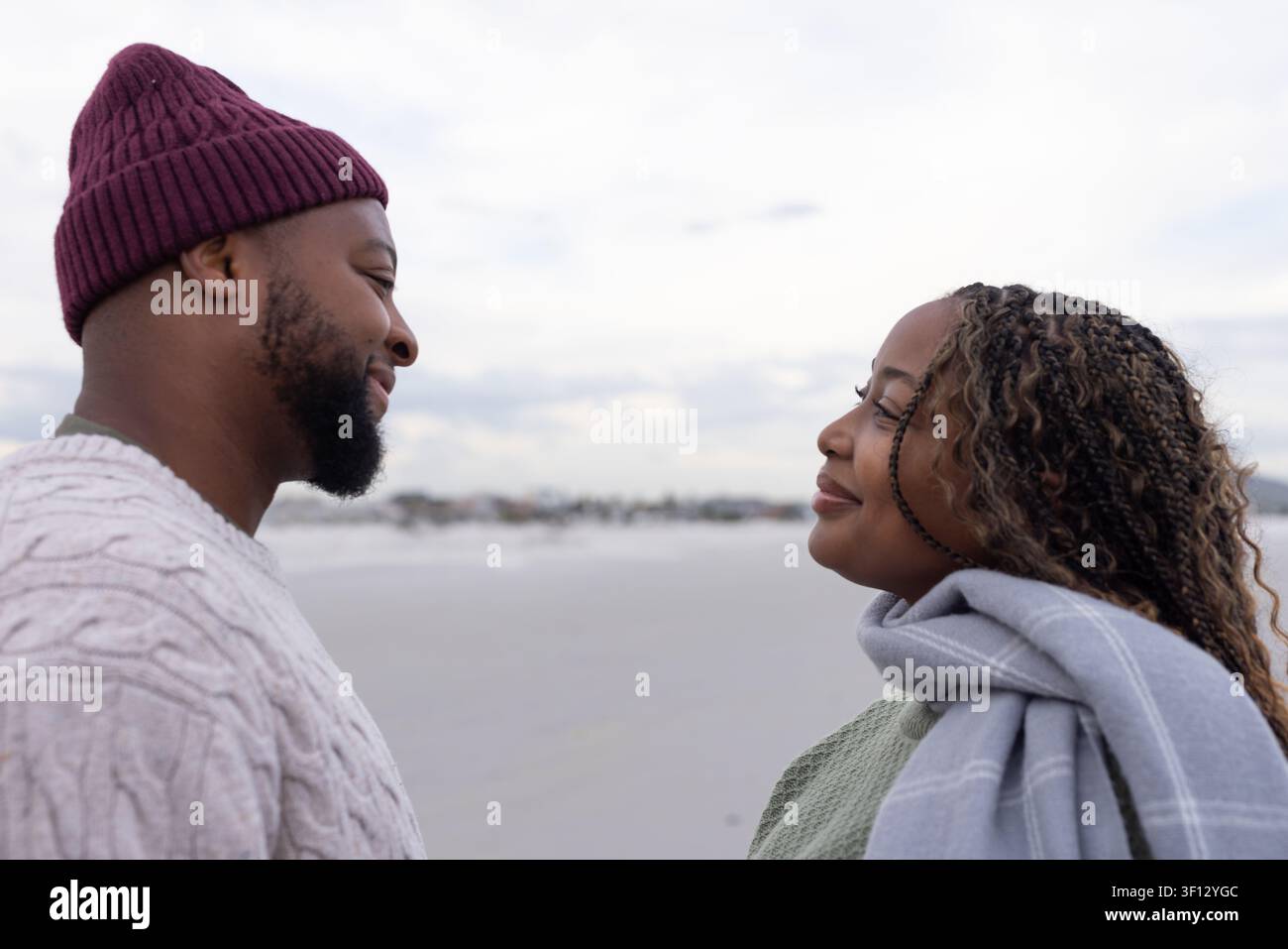 Coppia afroamericana in piedi uno di fronte all'altro su una spiaggia sabbiosa, indossa maglioni di lana e un beanie Foto Stock
