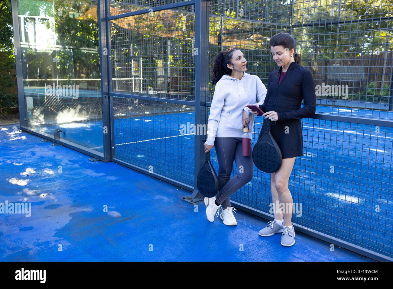 Diverse amiche in abbigliamento sportivo appoggiate a un campo blu con le racchette, che controllano il telefono, spazio di copia Foto Stock