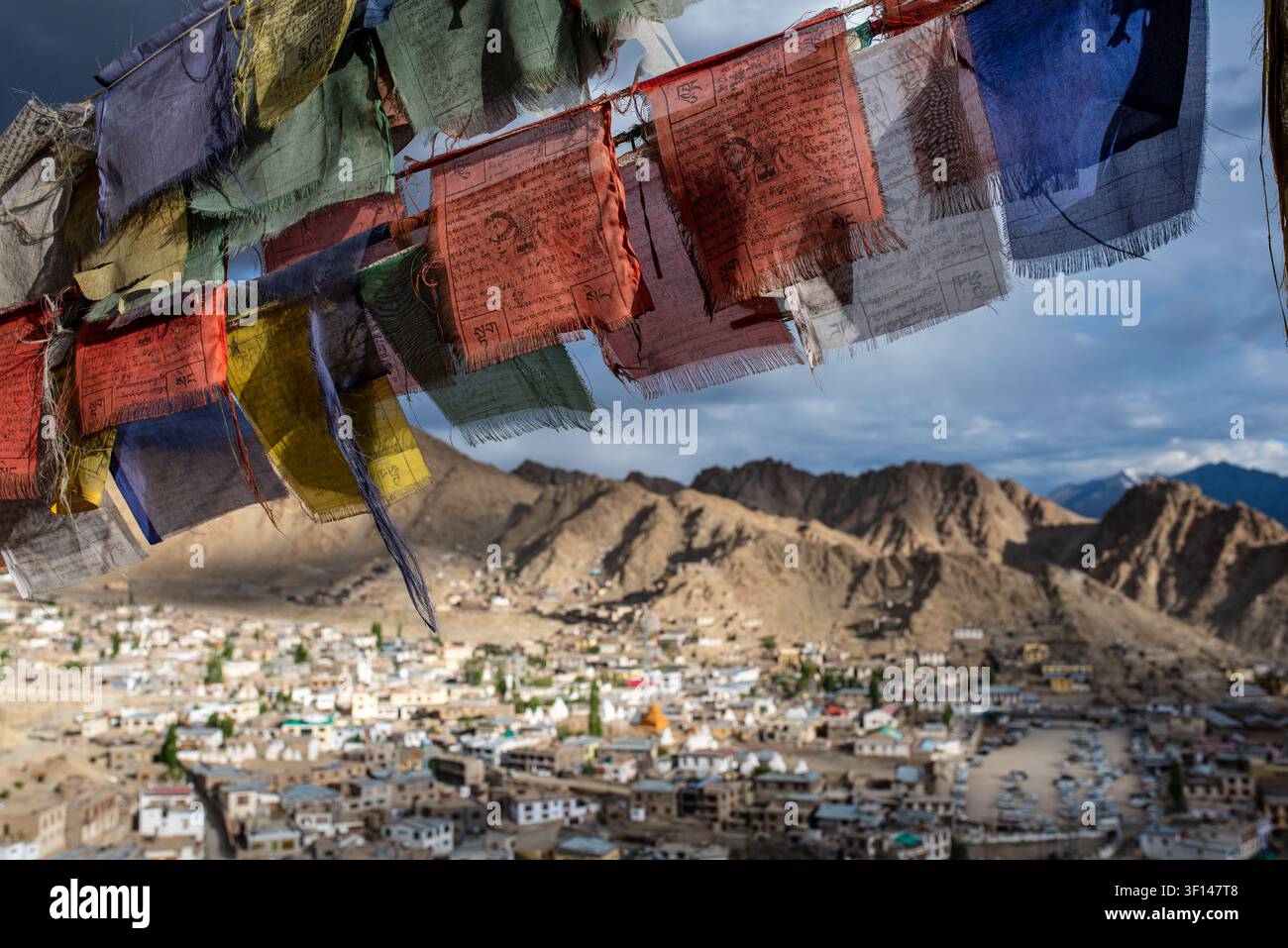 Bandiere di preghiera tibetane sfilacciate e colorate sventolano in primo piano, aprendosi su una vasta vista panoramica ad angolazione alta sulla città di Leh e il maestoso Barr Foto Stock