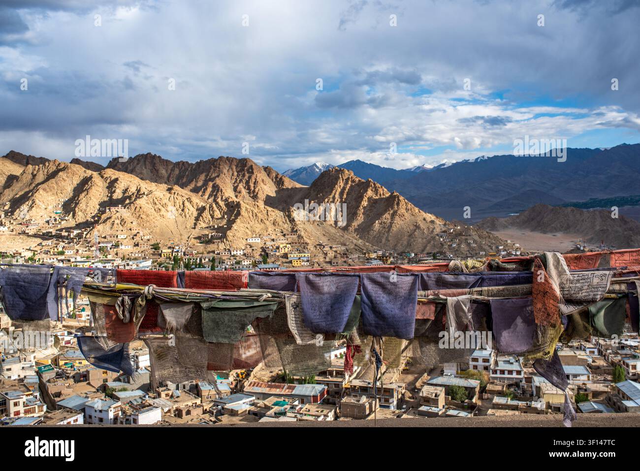 Bandiere di preghiera tibetane sfilacciate e colorate sventolano in primo piano, aprendosi su una vasta vista panoramica ad angolazione alta sulla città di Leh e il maestoso Barr Foto Stock