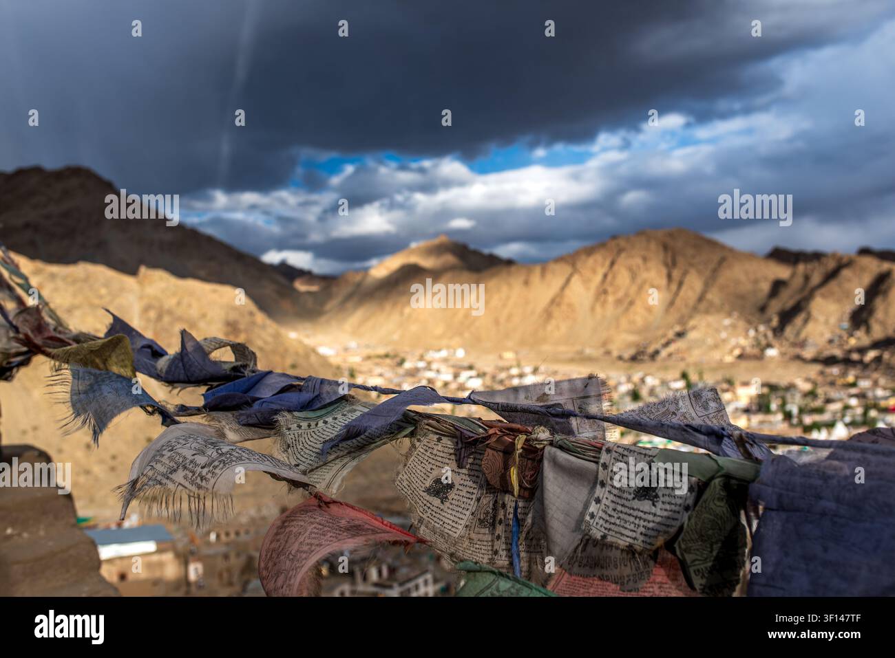 Bandiere di preghiera tibetane sfilacciate e colorate sventolano in primo piano, aprendosi su una vasta vista panoramica ad angolazione alta sulla città di Leh e il maestoso Barr Foto Stock