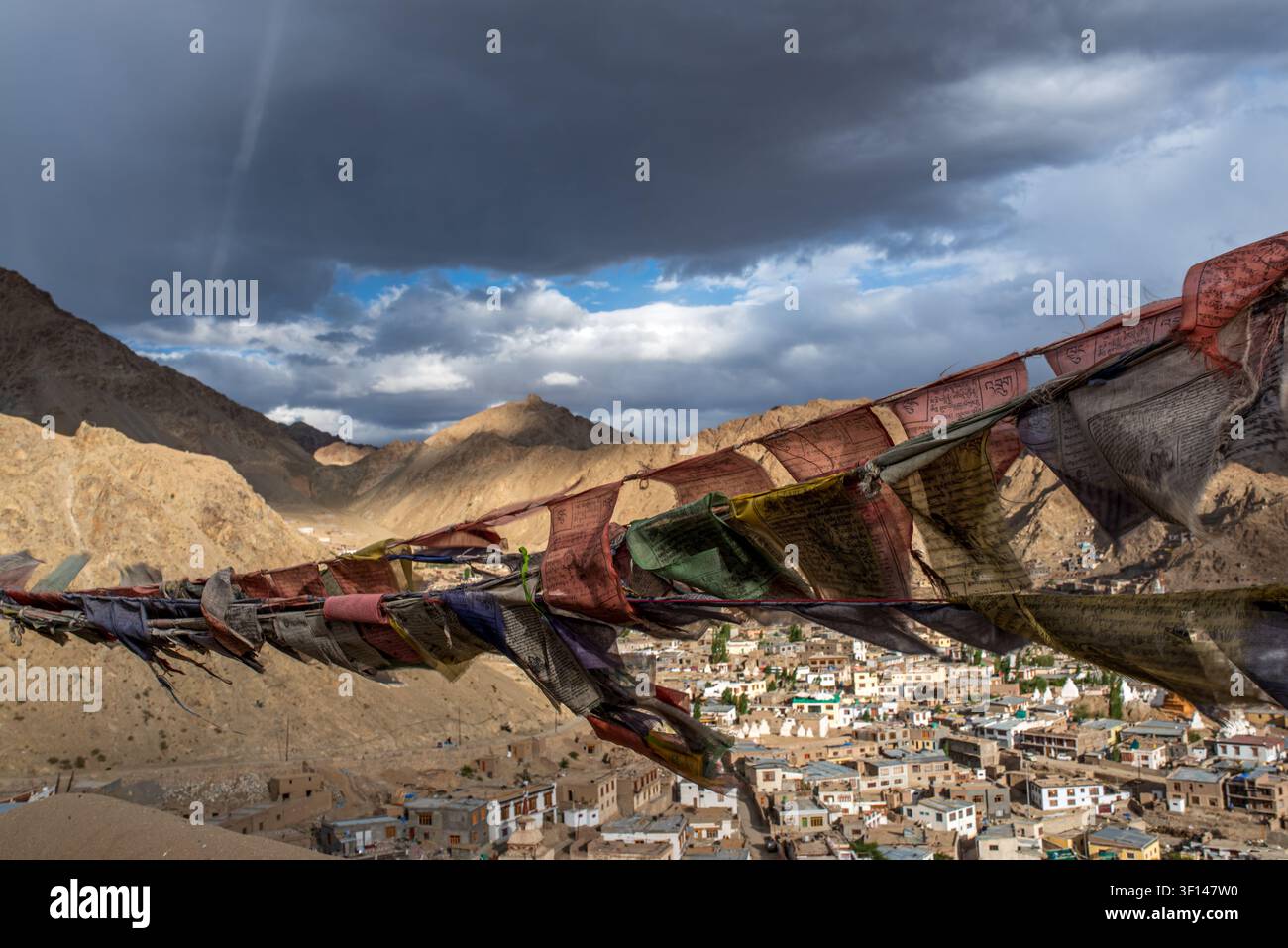 Bandiere di preghiera tibetane sfilacciate e colorate sventolano in primo piano, aprendosi su una vasta vista panoramica ad angolazione alta sulla città di Leh e il maestoso Barr Foto Stock