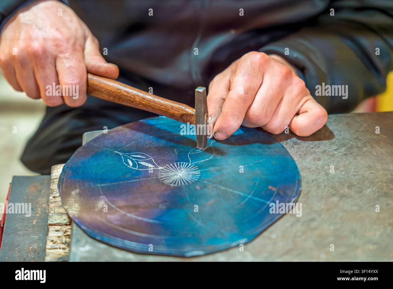 Un primo piano delle mani di un artigiano che incide un tradizionale motivo damascato su un disco metallico blu, utilizzando uno scalpello e un martello, cattura un secolo Foto Stock
