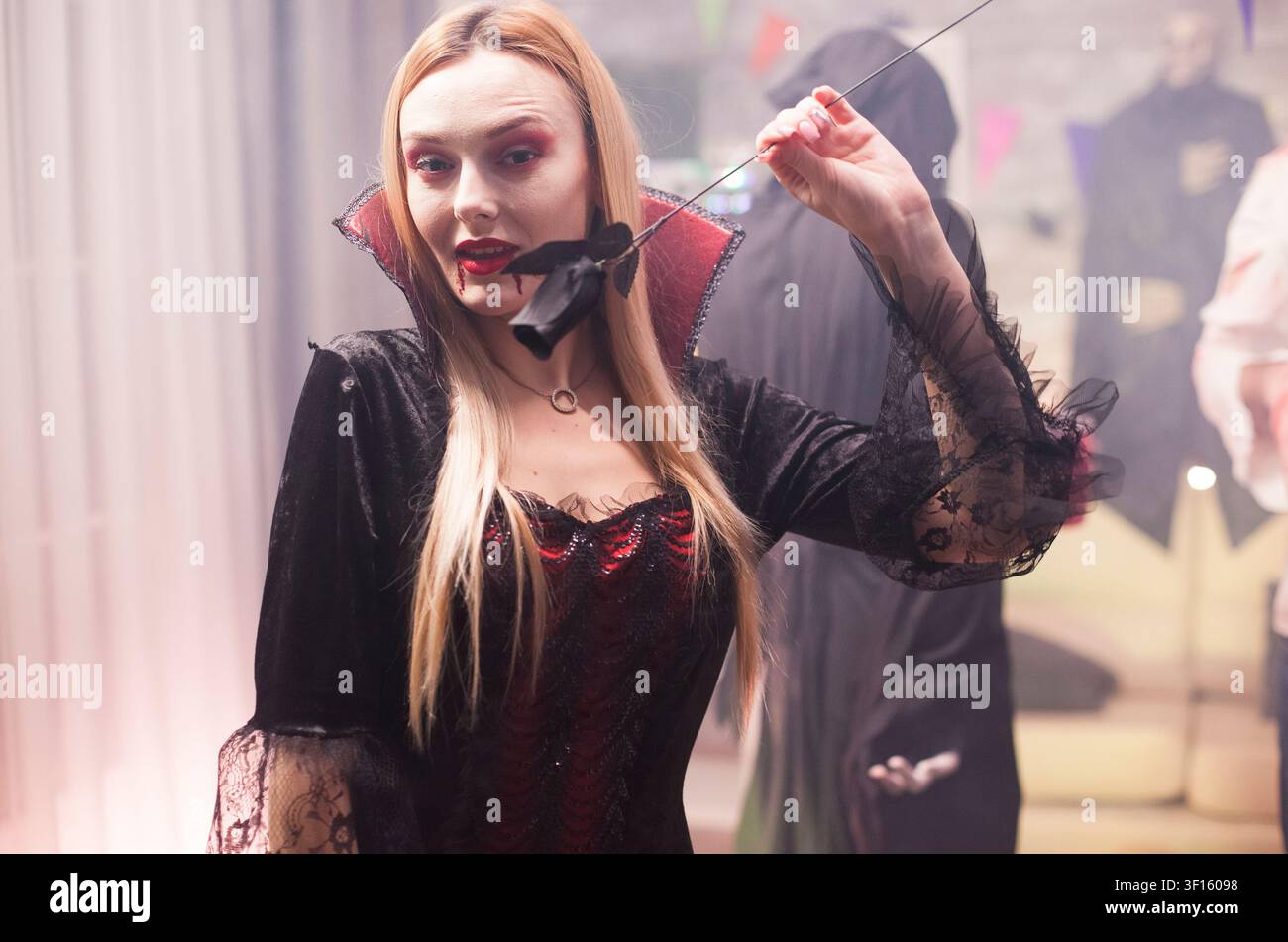 Giovane donna vestita da vampiro spaventoso celebra Halloween con un gruppo di persone in costume, mostrando uno stile horror giocoso, attività di gruppo tra amici. Foto Stock