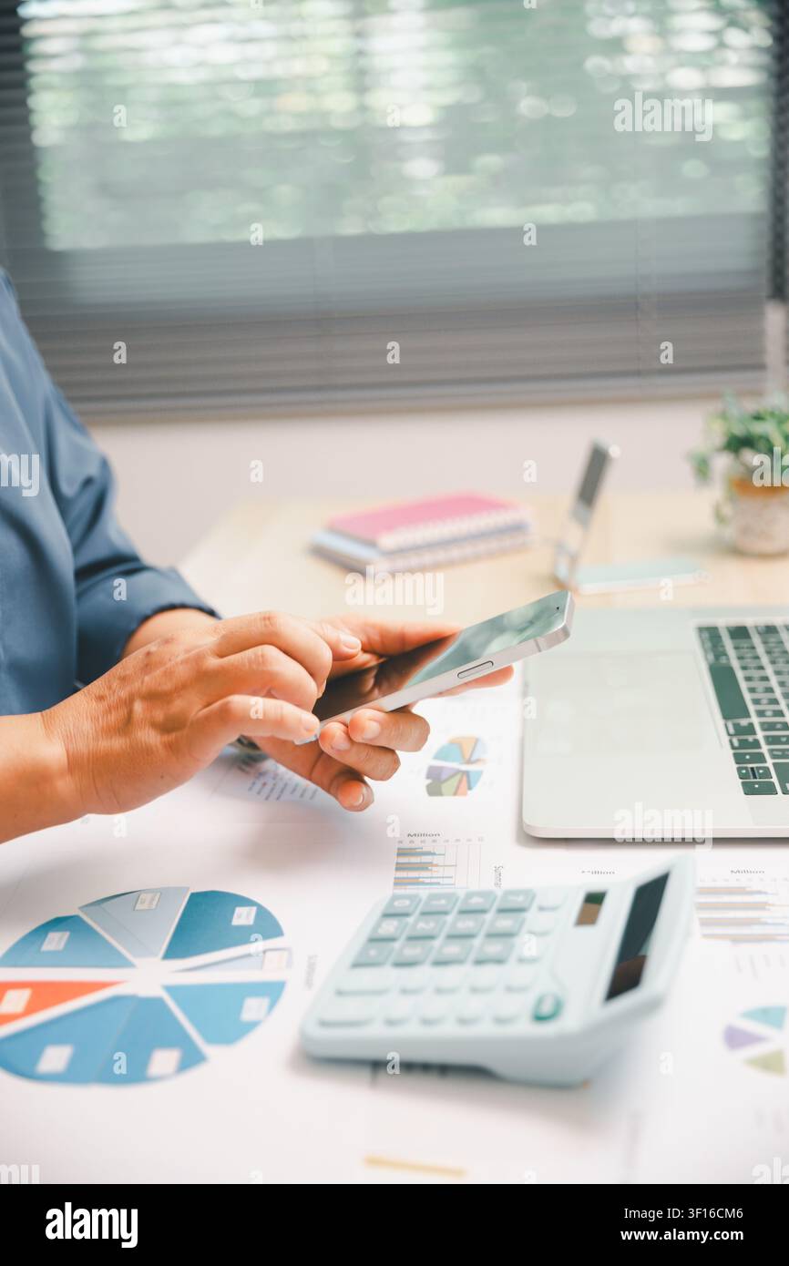 Mani professionali che utilizzano uno smartphone accanto a un laptop, una calcolatrice e grafici finanziari sulla scrivania dell'ufficio, mostrando attività mobile, revisione contabile, dati Foto Stock