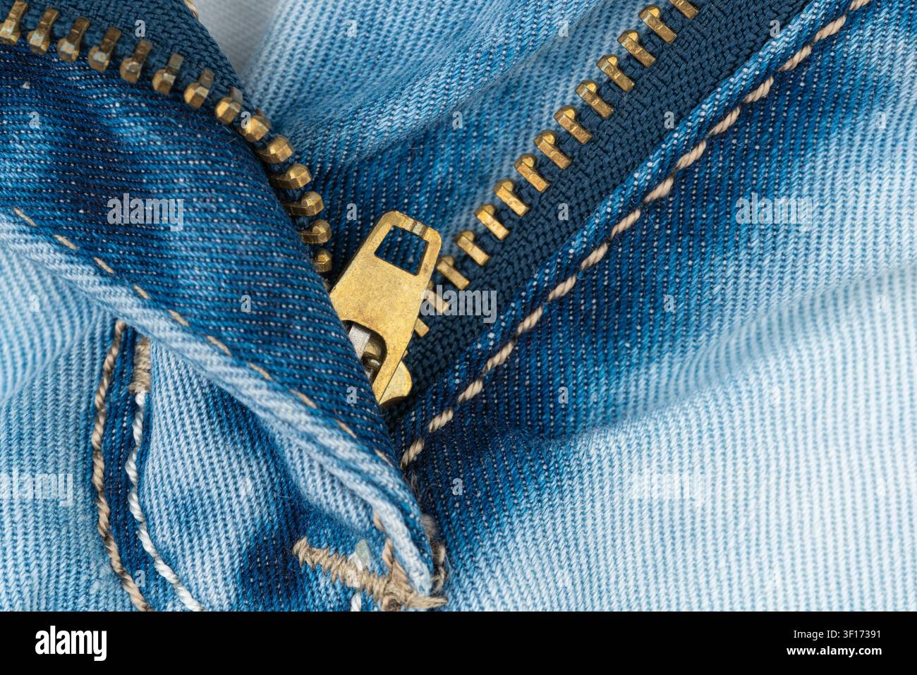 Estremo primo piano di una cerniera aperta su jeans blu. Texture dettagliata del tessuto denim con chiusura in ottone. La fotografia macro mostra le pieghe di cucitura nel materiale denim Foto Stock