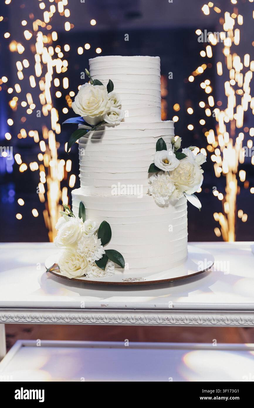 Torta nuziale elegante con glassa bianca morbida e fiori. Fiori delicati adornano ogni piano con verde. Luci tremolanti creano un bagliore romantico beh Foto Stock