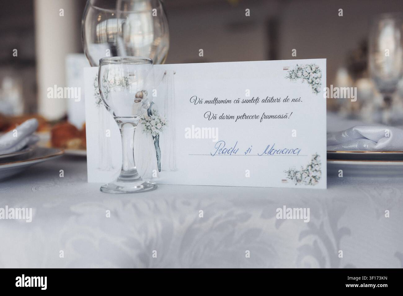 Impostazione elegante del tavolo con bicchieri e biglietto floreale. Una nota manoscritta suggerisce un matrimonio o una celebrazione. Una tovaglia bianca aggiunge uno sfondo raffinato e pulito. Foto Stock