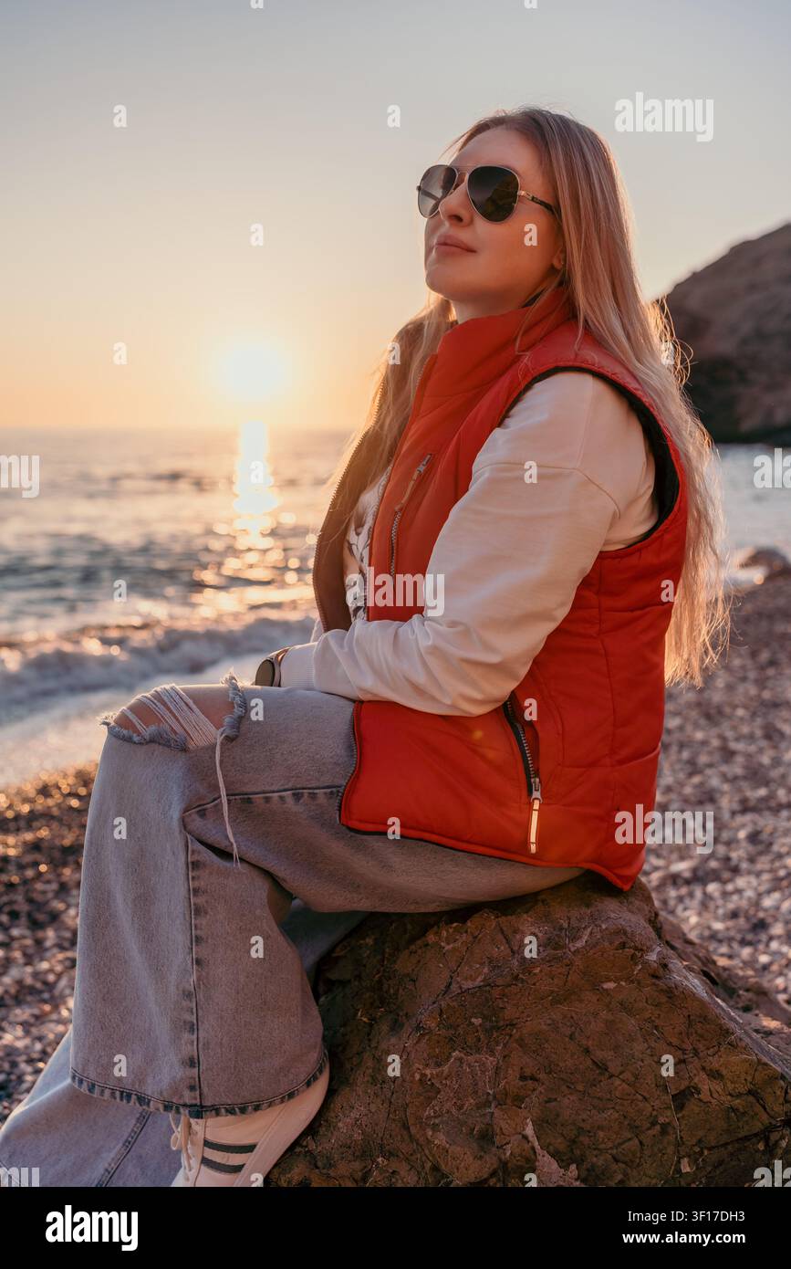 Donna seduta su una roccia in spiaggia al tramonto, indossa un giubbotto rosso e occhiali da sole mentre gode dell'ora dorata sulle onde dell'oceano Foto Stock