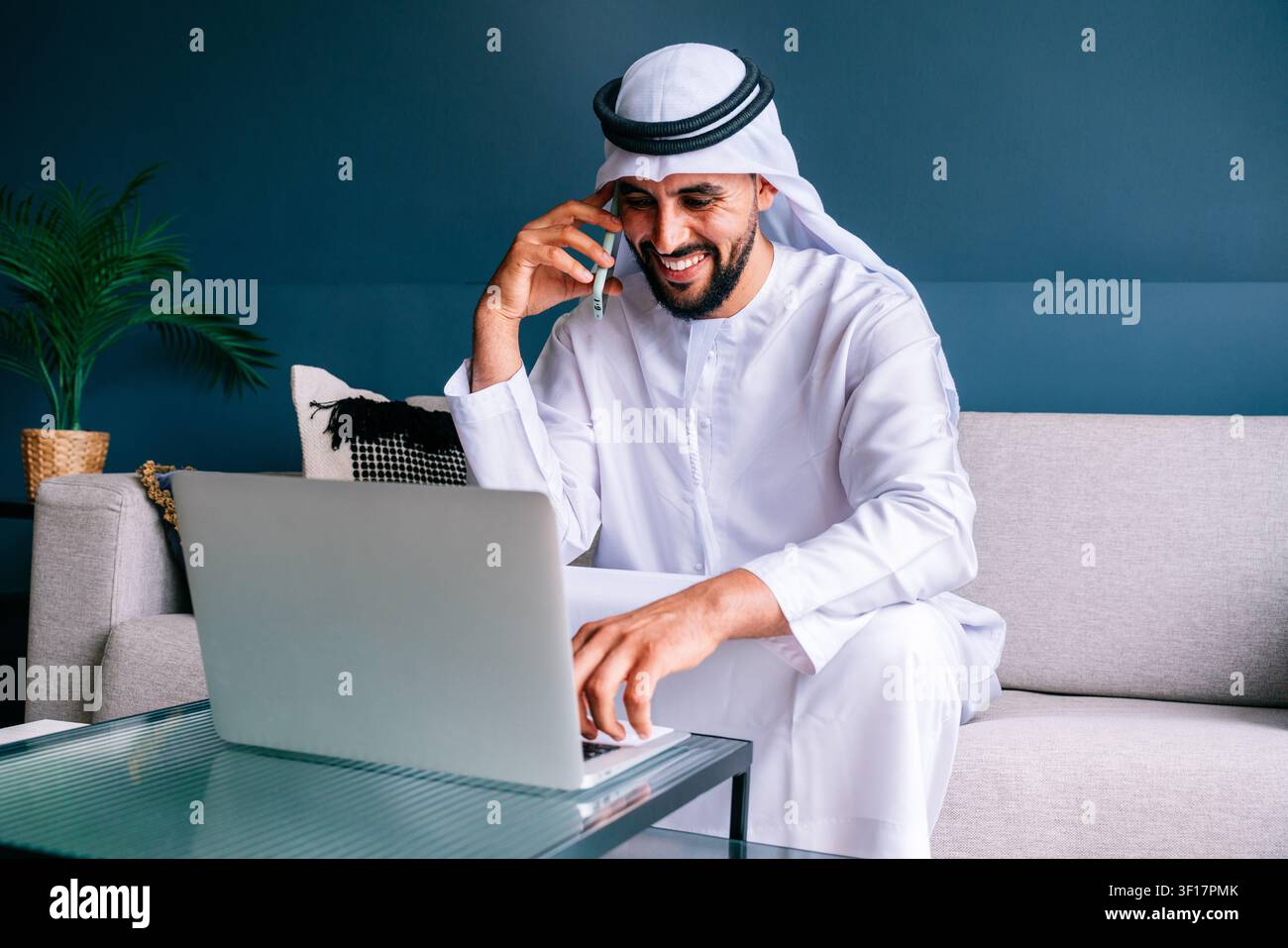 Uomo emiratino che indossa kandura e ghutra, felicemente impegnato nel lavoro a distanza, parlando al telefono cellulare e digitando su un laptop dal suo soggiorno moderno Foto Stock