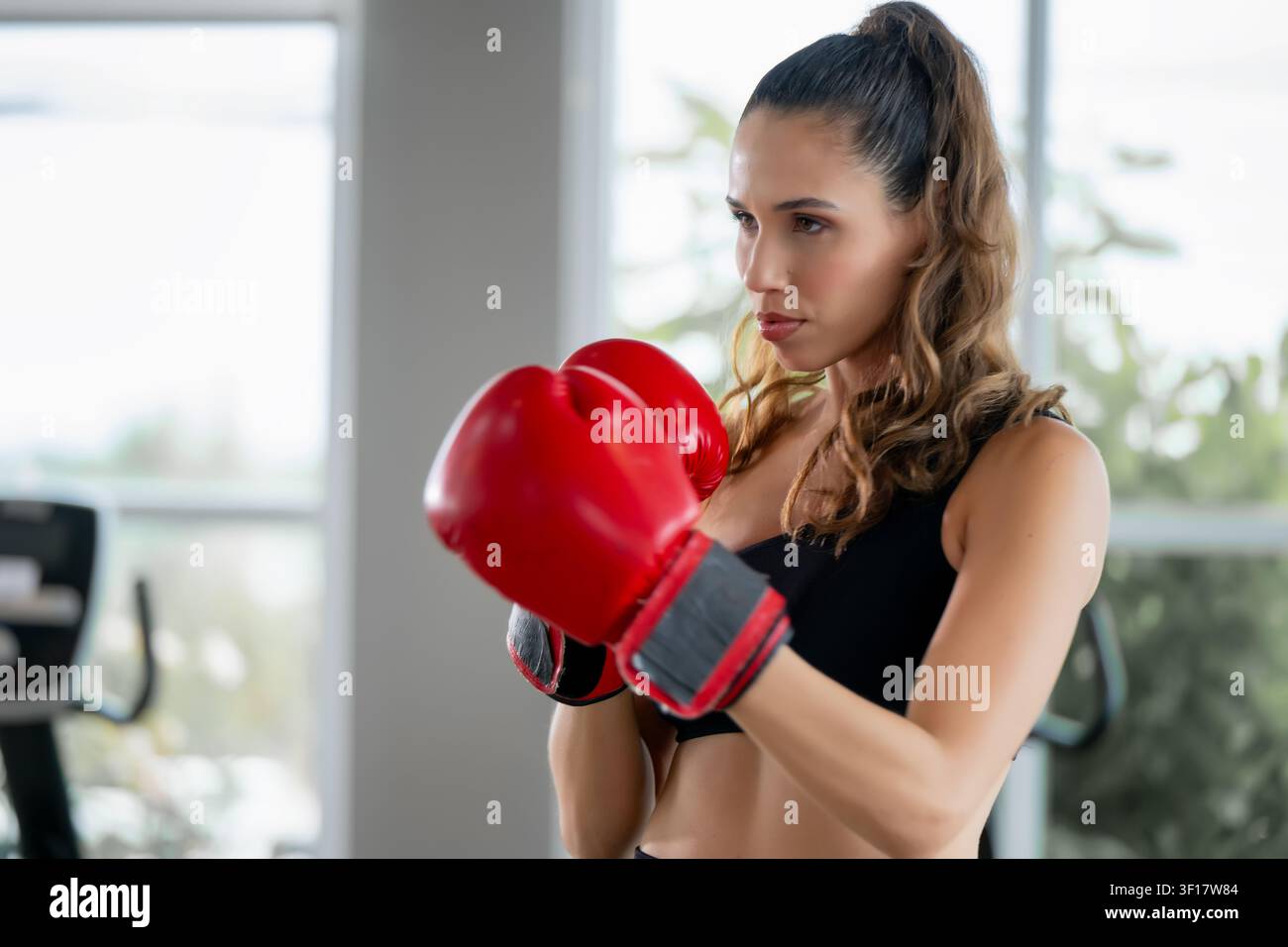 Ritratto di una giovane donna sicura di sé che indossa guantoni da boxe rossi in una palestra, con uno sguardo determinato e concentrato mentre pratica la boxe e si allena. Sce Foto Stock
