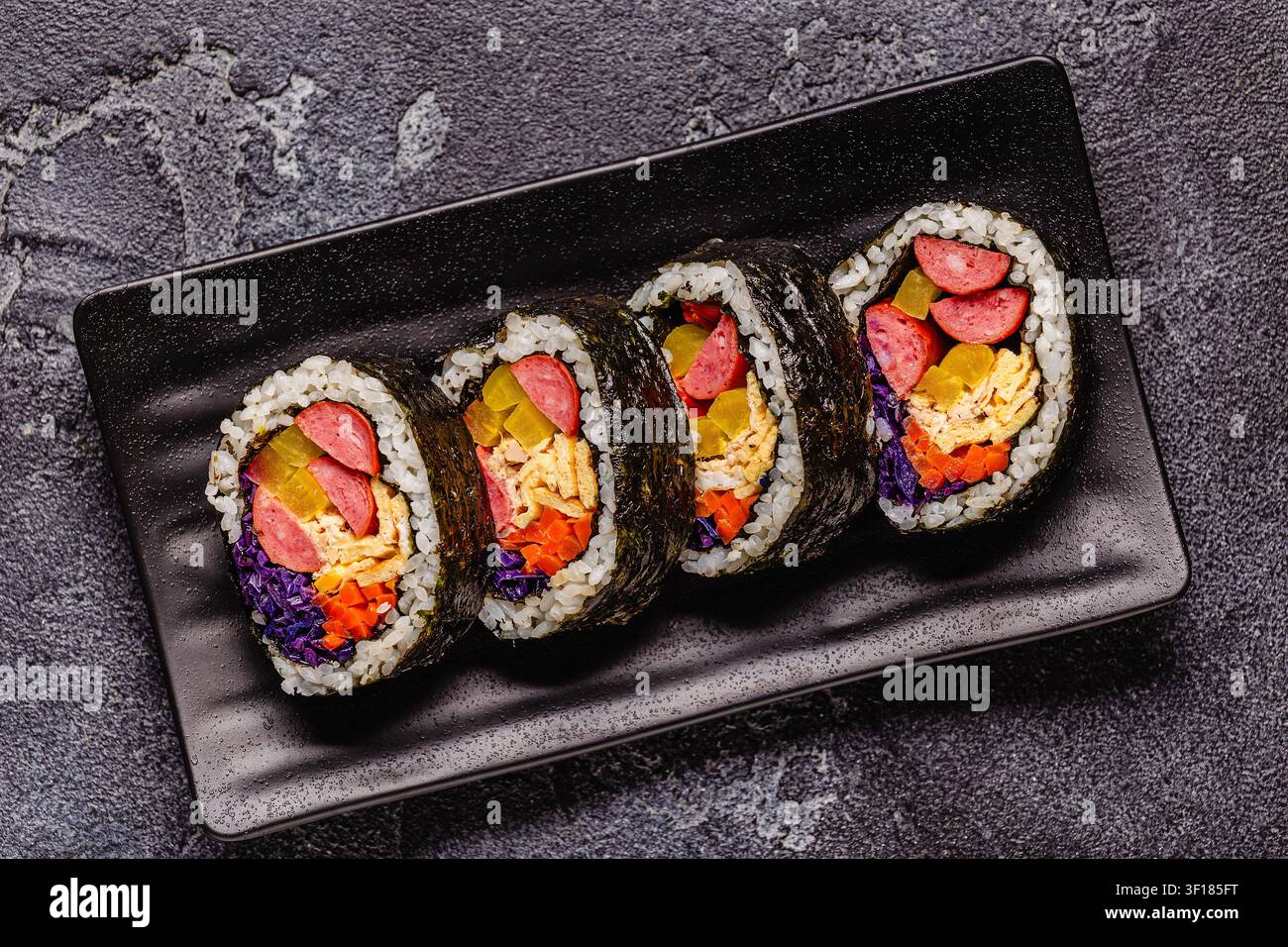 Un piatto nero pieno di kimbap (o gimbap) con ripieni colorati. Gli ingredienti includono salsiccia, cetrioli, uovo e verdure. Foto Stock