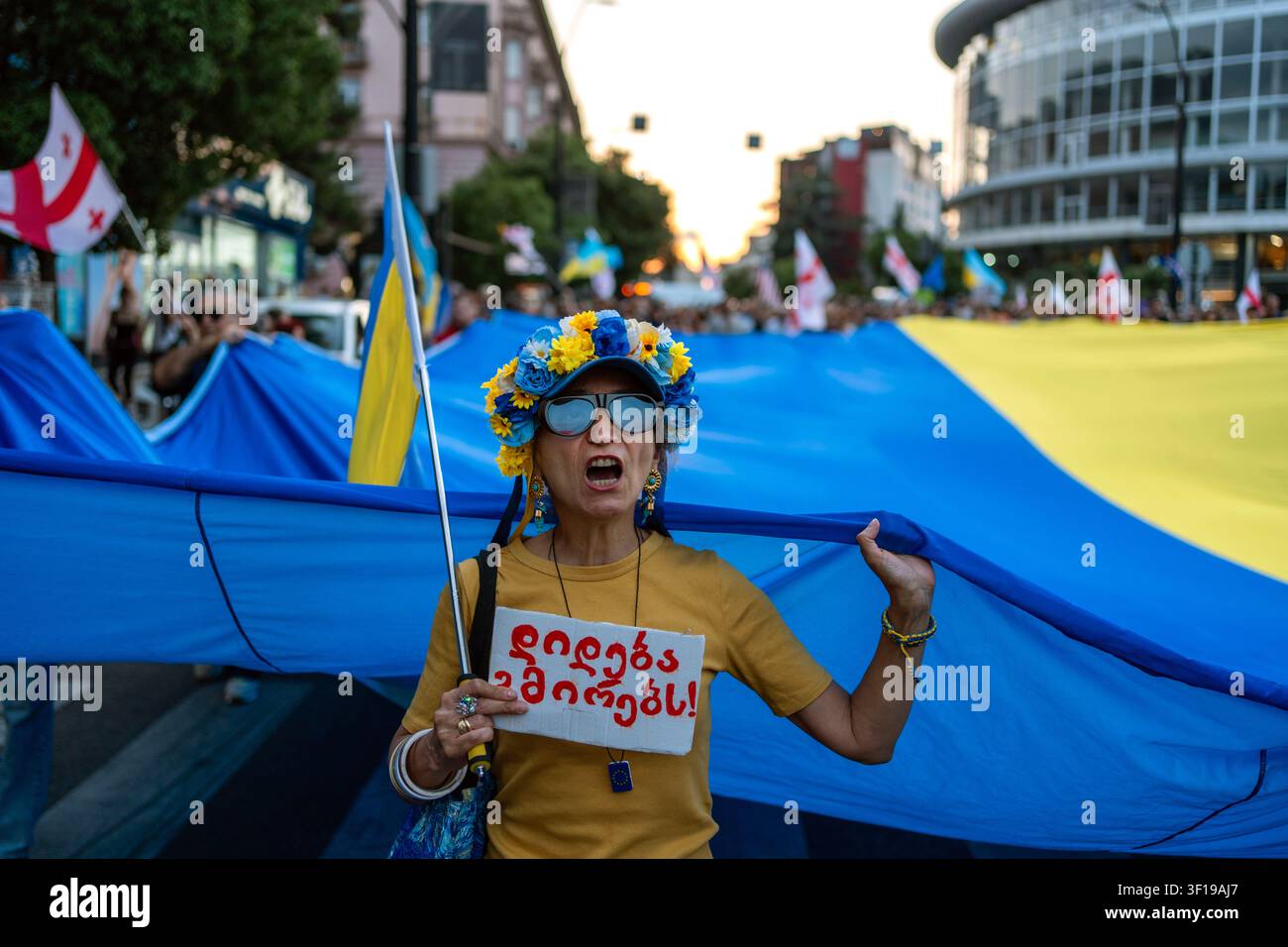 Tbilisi, Georgia, 11 luglio 2026. Una donna canta mentre lei e altri manifestanti portano una grande bandiera ucraina per celebrare il 591º giorno della manifestazione antigovernativa. Foto Stock