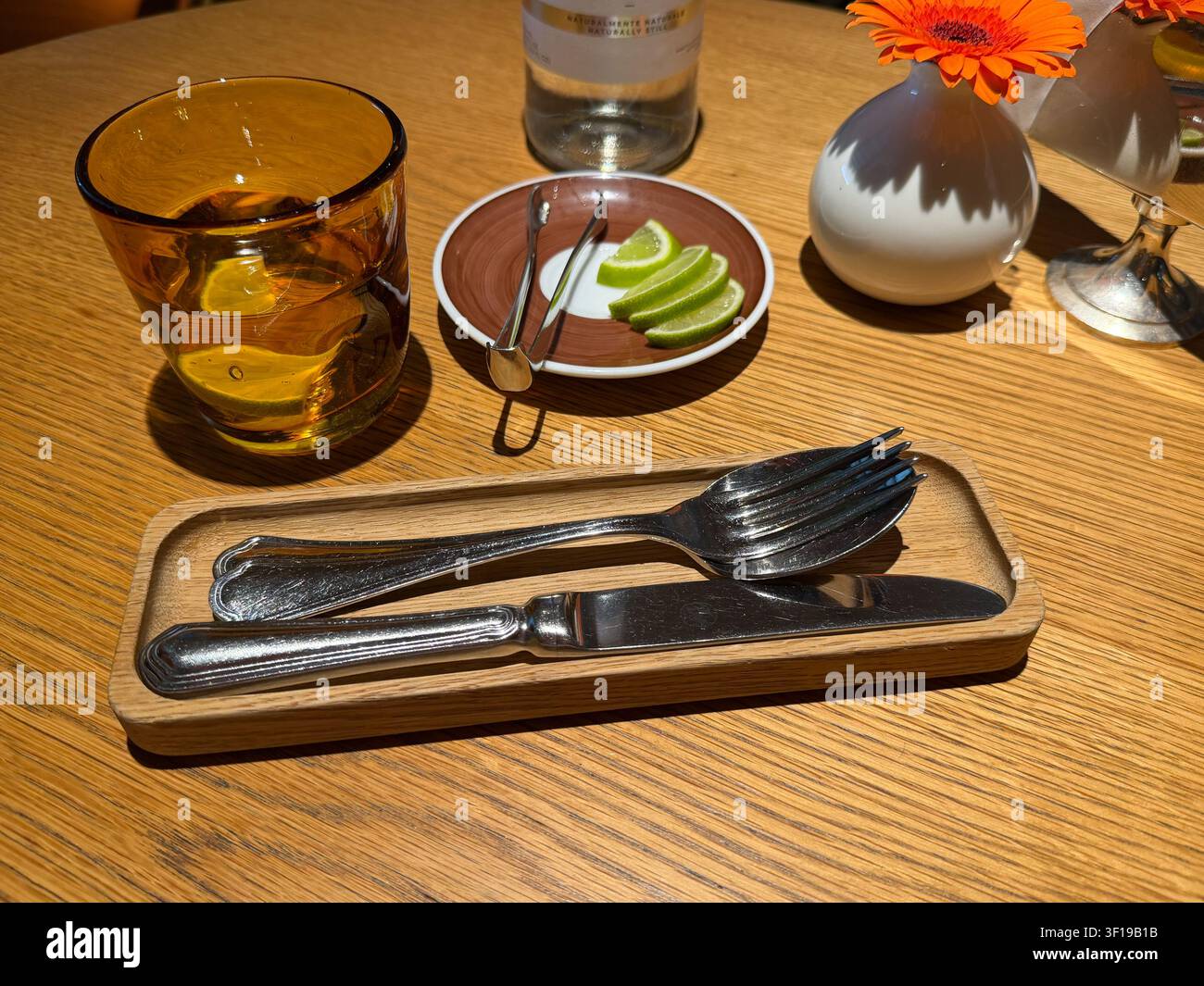 Apparecchiatura da tavolo con posate, bicchiere d'acqua ambrato, spicchi di lime e vaso di fiori su tavolo di legno in un caffè moderno. Interno di ristorante, cena, ospitalità Foto Stock