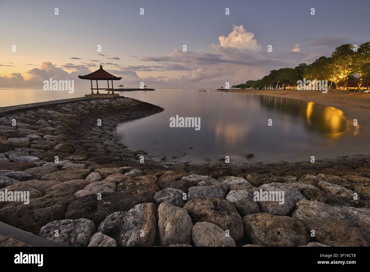 Vista serena del mattino da un molo di pietra su una baia tranquilla a Sanur Beach all'alba, con le luci dei resort che si riflettono nell'acqua immobile a Bali Foto Stock