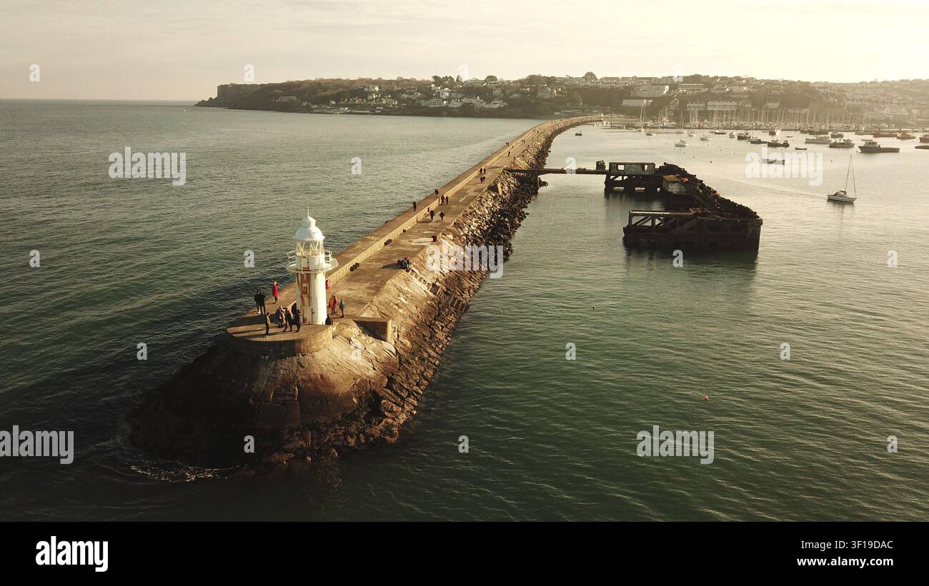 Un lungo molo con un faro si estende nell'oceano con barche e una città sullo sfondo, Brixham, Devon, Regno Unito Foto Stock