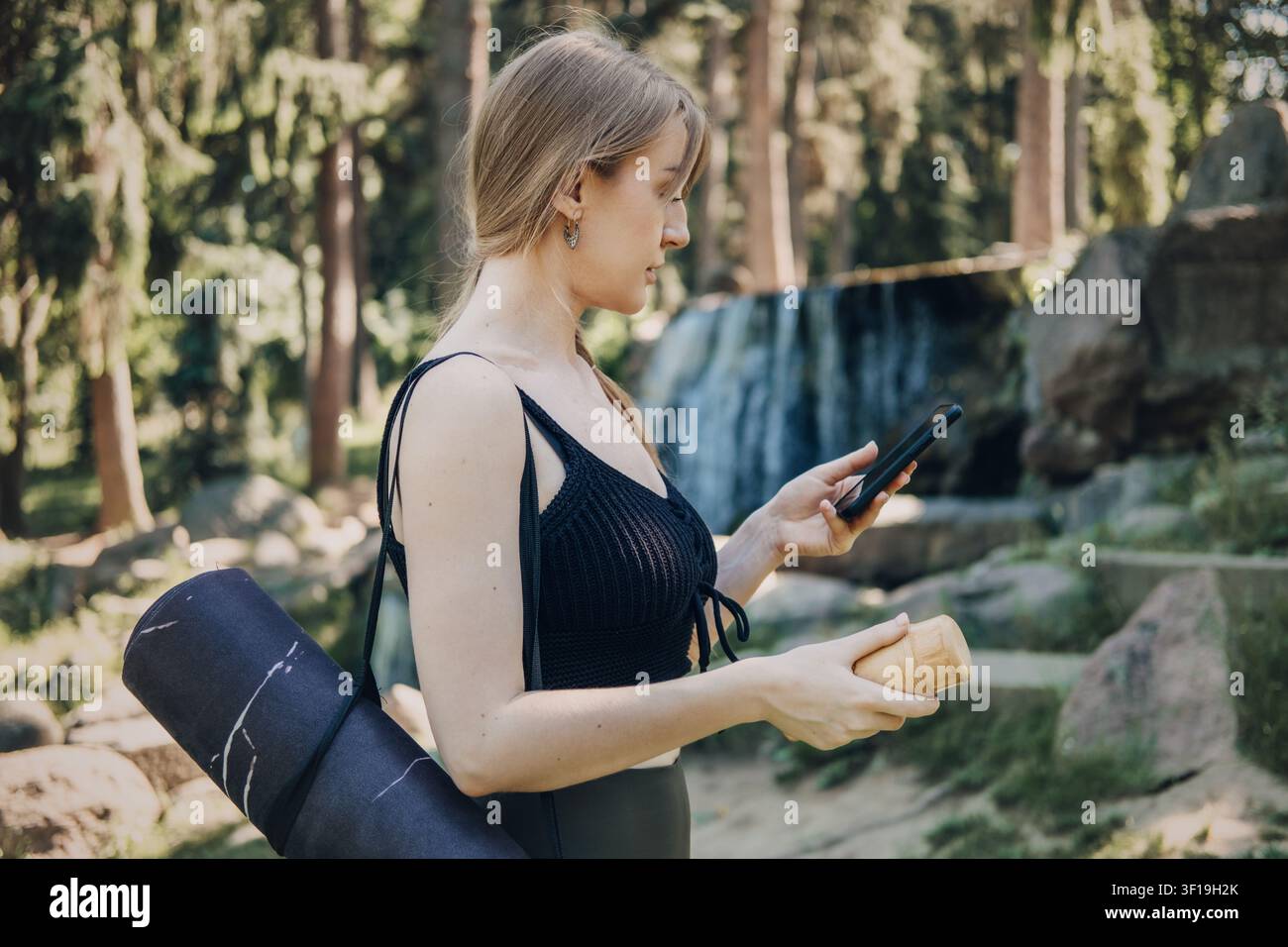 Donna tiene uno smartphone e una borraccia mentre porta un tappetino da esercizio arrotolato all'aperto vicino a una cascata. Alternativa alla palestra, allenamento basato sulla natura, nessun Foto Stock