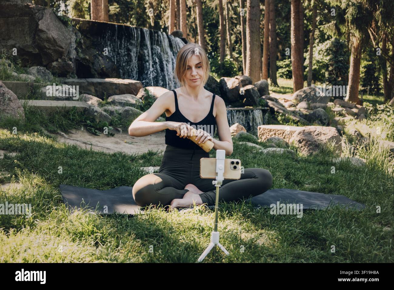 Donna apre una bottiglia d'acqua riutilizzabile mentre è seduta su un tappeto davanti a uno smartphone su un treppiede. Insegnamento di fitness a distanza, corso di benessere digitale, consapevolezza Foto Stock