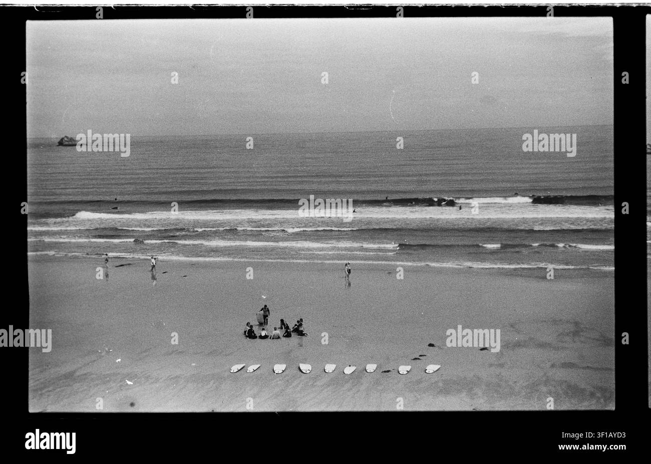 I surfisti sono seduti insieme dietro una fila ordinata di tavole mentre un'onda più grande si infrange attraverso il paesaggio marino monocromo aperto. Foto Stock