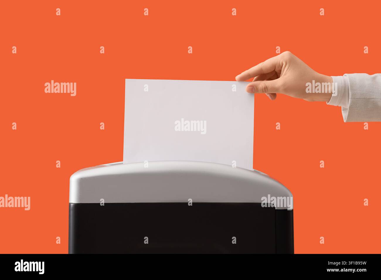 Mano che inserisce un foglio di carta bianca vuoto in un trituratore di documenti per la distruzione e la sicurezza dei dati su uno sfondo arancione Foto Stock