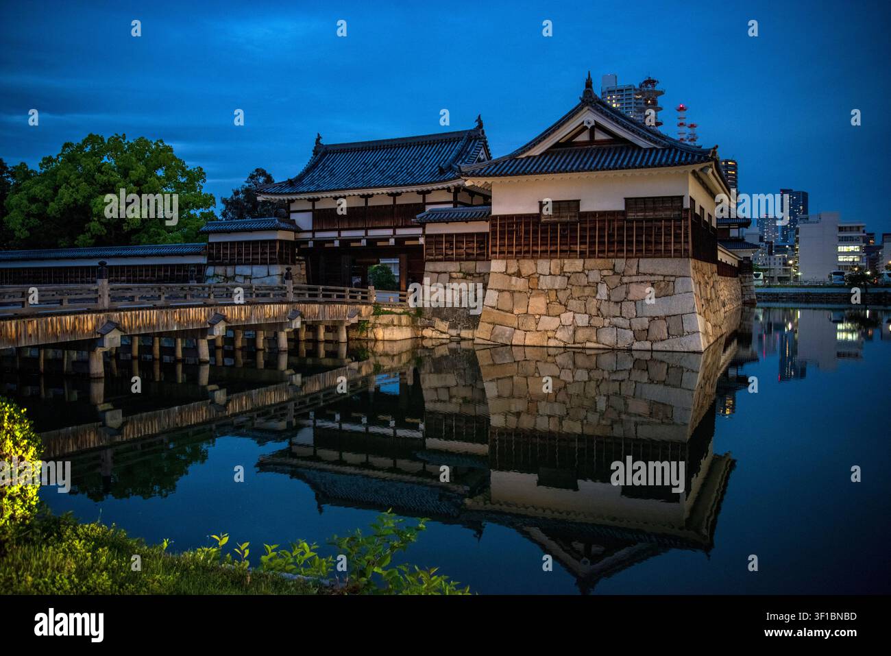 Il ponte di legno ricostruito storico Gomonbashi e il cancello Omote-gomon del castello di Hiroshima si riflettono nell'acqua calma del fossato al crepuscolo a Hiroshima Foto Stock