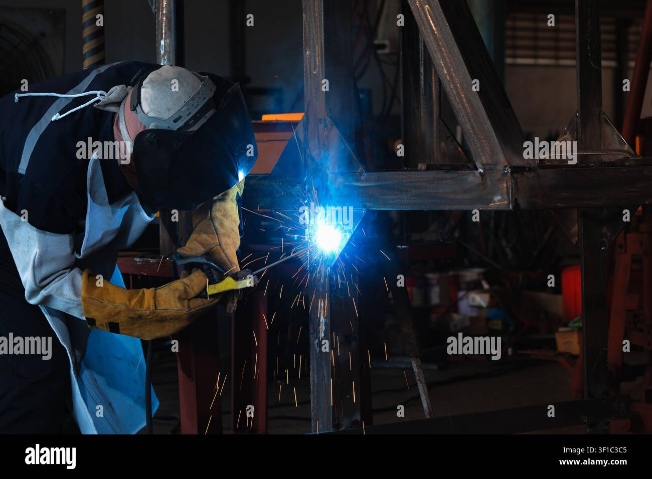 Il saldatore indossa un casco e guanti. Scintille blu brillanti volano dalla torcia saldante. Si concentra sull'unire travi metalliche. Lavoro industriale in fabbrica. Abile c Foto Stock