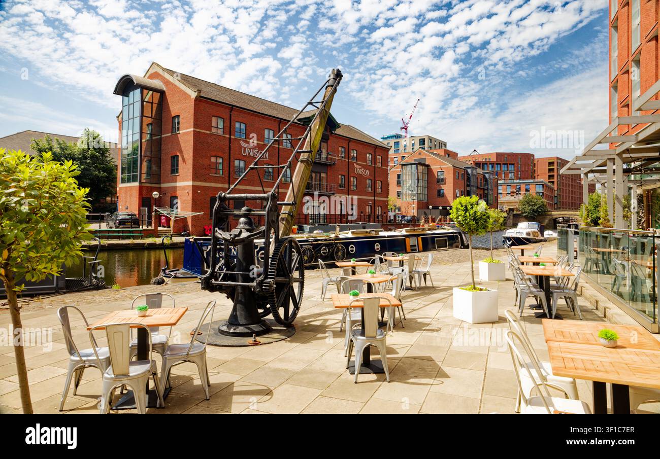 Una vecchia gru di sollevamento all'interno dell'area di seduta esterna di un bar sul lato del canale con chiatte ed edifici per uffici al Granary Wharf, Leeds, West Yorkshire. Foto Stock