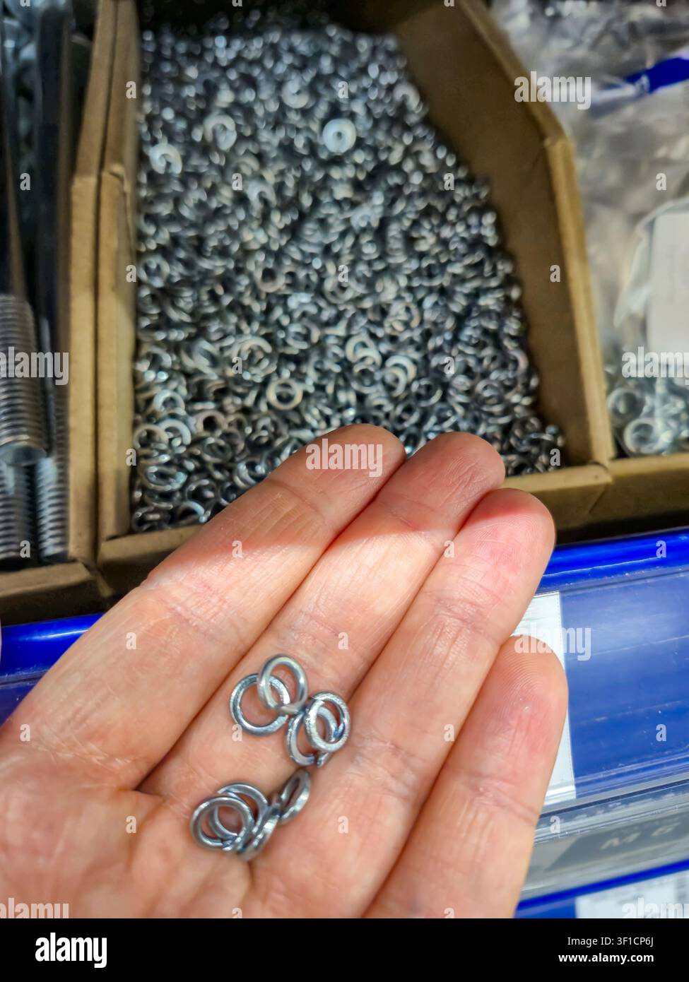 Il palmo di una persona mostra una collezione di piccole rondelle metalliche lucide, con uno sfondo sfocato di molti altri elementi di fissaggio in contenitori Foto Stock