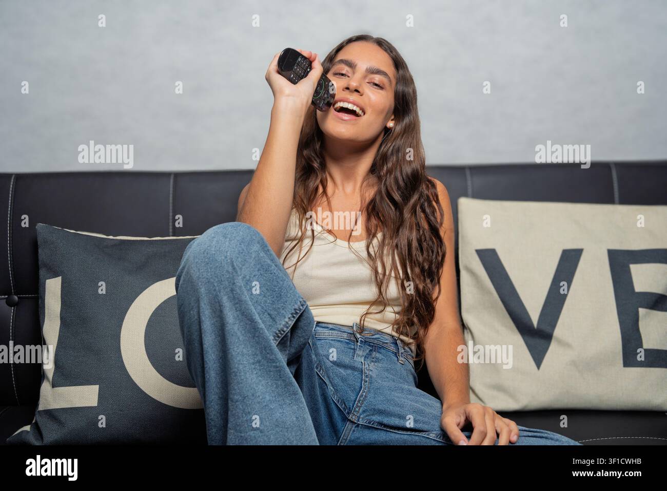 Giovane donna che canta felicemente in un telecomando mentre si rilassa su un divano a casa Foto Stock