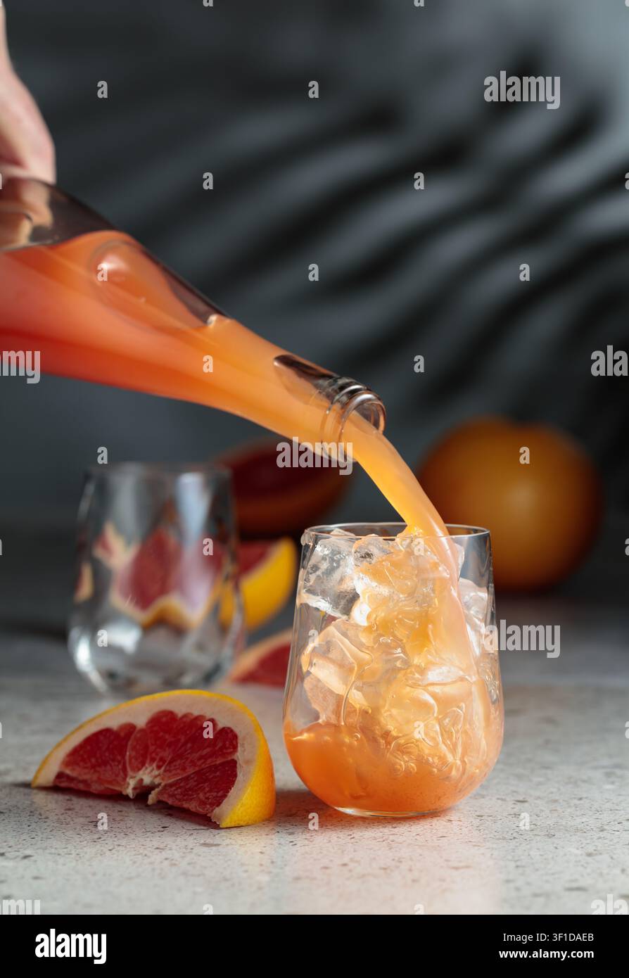 Il succo di pompelmo viene versato in un bicchiere con ghiaccio. Bere in un bicchiere congelato e frutta fresca su un tavolo di pietra. Sullo sfondo, le ombre del palmo leav Foto Stock