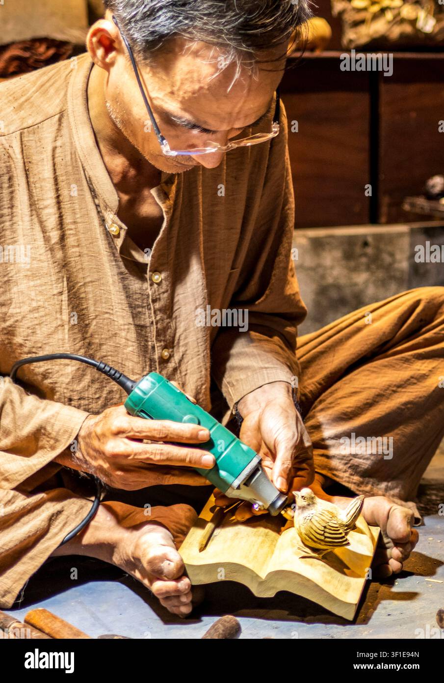 Hoi An, Vietnam – 28 giugno 2025: Di notte, un uomo seduto fuori dal suo negozio e sostiene il legno con i piedi, modella con cura la bella opera d’arte con Foto Stock