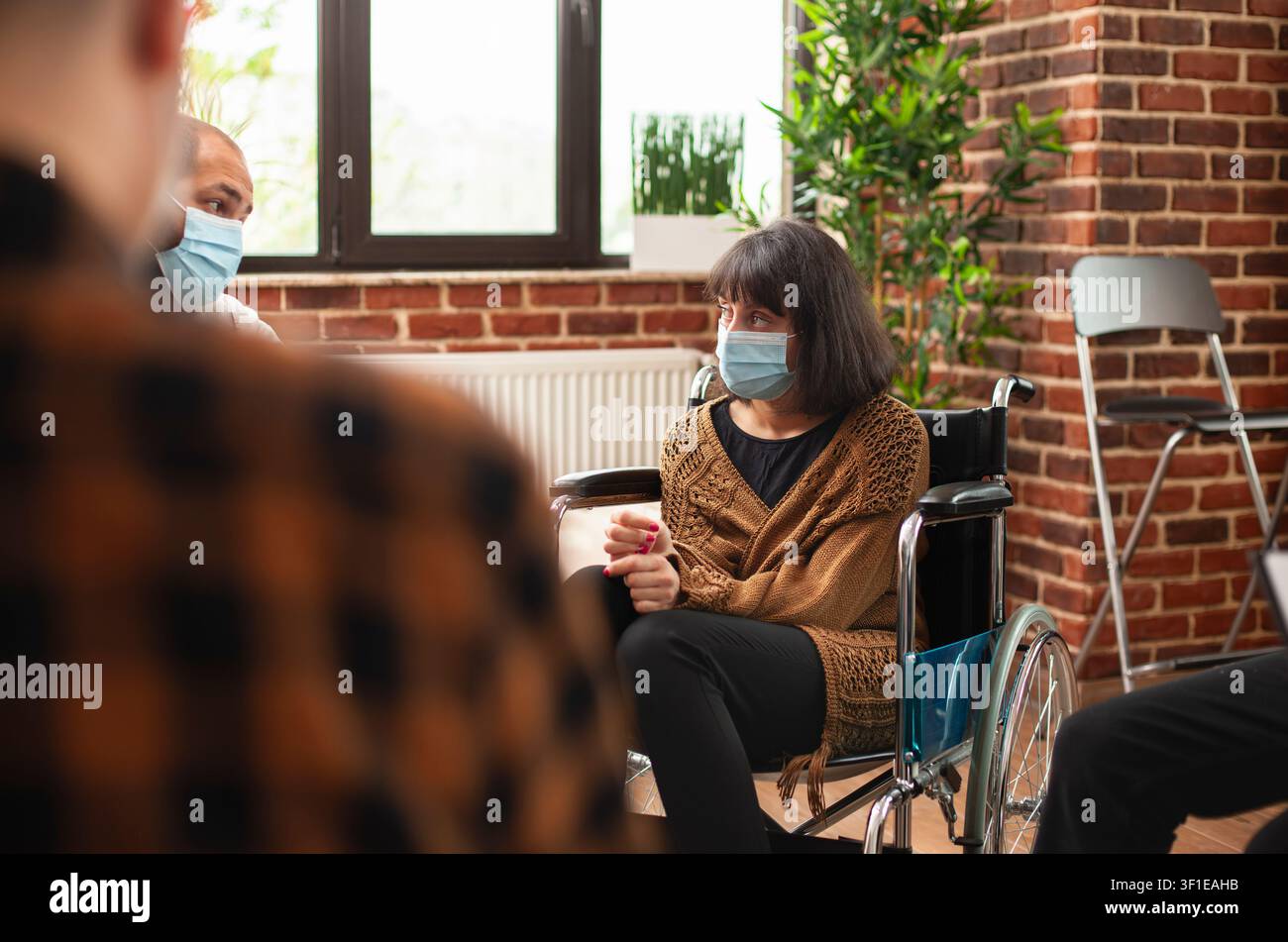 Giovane utente in sedia a rotelle ascolta durante una terapia di gruppo mascherata, mostrando consulenza per disabili, supporto riabilitativo e condivisione emotiva in un ufficio di mattoni. Foto Stock