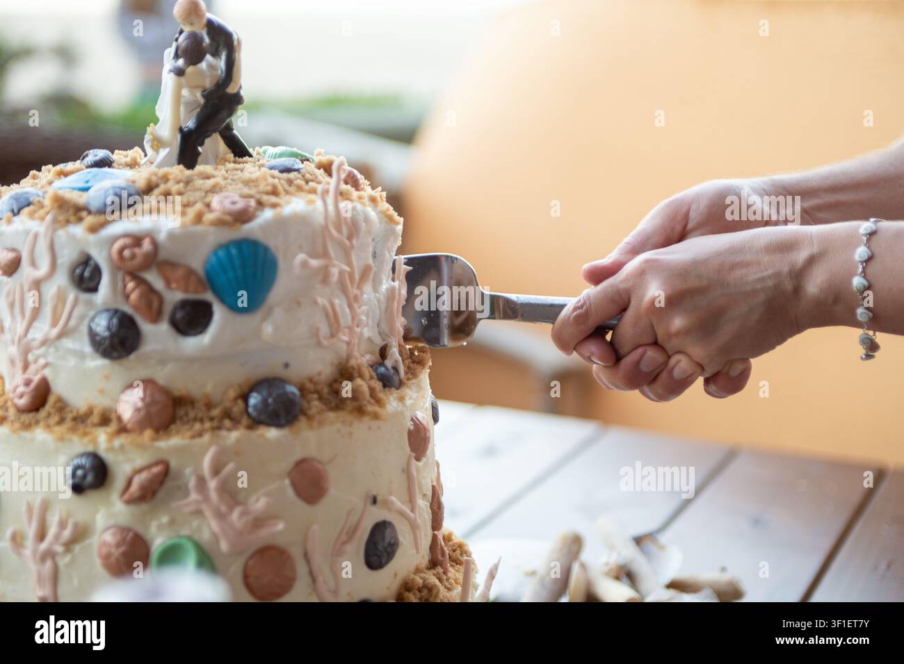 Le mani di una coppia di sposi novelli tagliano una torta nuziale a più piani decorata con conchiglie commestibili e sabbia. Foto Stock