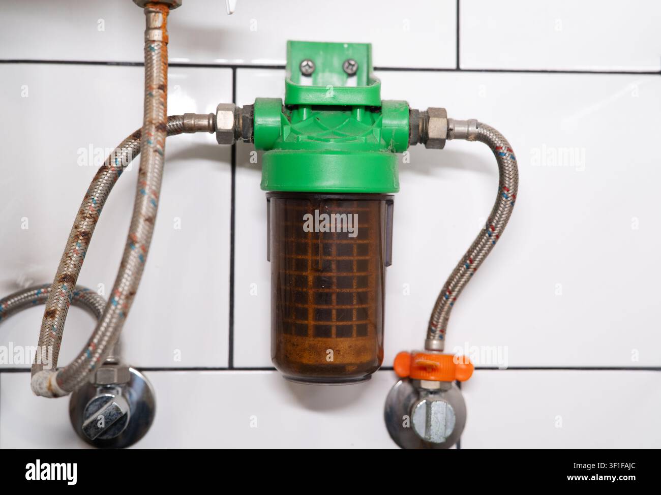 Alloggiamento a rete per filtro dell'acqua sporco collegato con tubi intrecciati arrugginiti su uno sfondo di pareti piastrellate bianche. Foto Stock