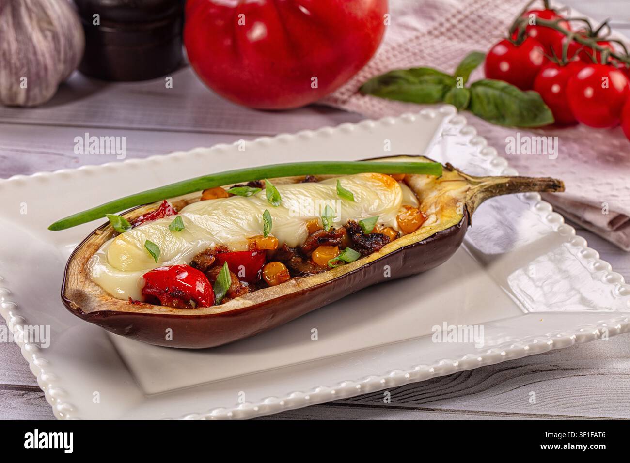 Mezza melanzana al forno ripiena di verdure miste e formaggio fuso, guarnita con cipolla verde. Servita su piatto di ceramica bianca con basilico al pomodoro sullo sfondo Foto Stock