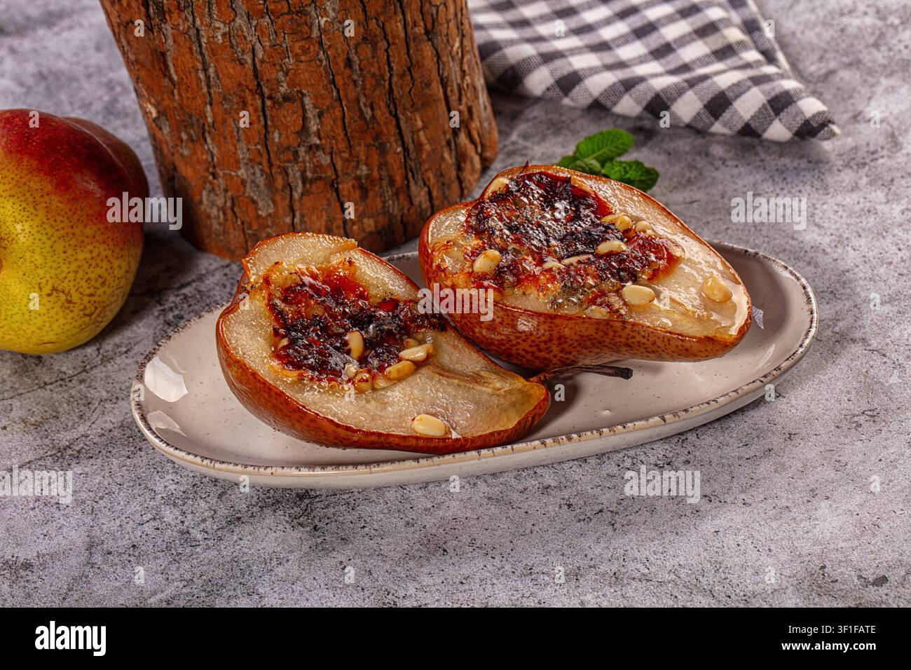 Mezze pere cotte con marmellata e pinoli su piatto di ceramica, pera intera e tronco di legno nelle vicinanze, fotografia alimentare rustica con luce calda e atmosfera autunnale Foto Stock